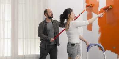 Family using orange color to paint wall with roller and paintbrush during home renovation. Painting with new color to redecorate apartment room, interior renovation and house improvement.