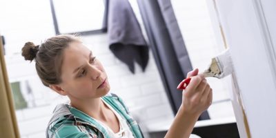 Home repairs. A focused woman paints in white the entrance to the bathroom with a brush and can, which she holds in her hands. Close-up.